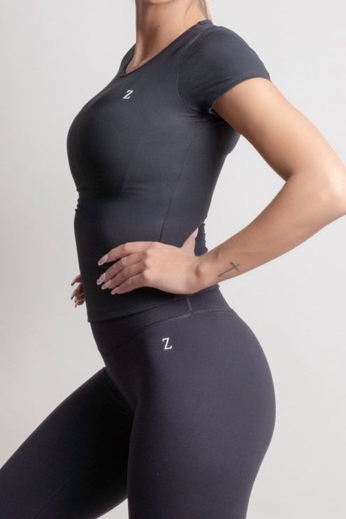 Woman wearing a NOIR t-shirt Black athletic outfit with white sneakers on a light gray background