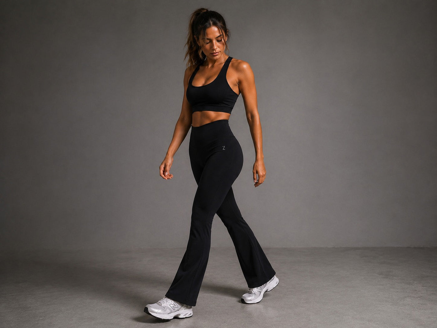 A woman modeling a black athletic set consisting of a sports bra and high-waisted flared leggings, walking against a neutral gray studio background.