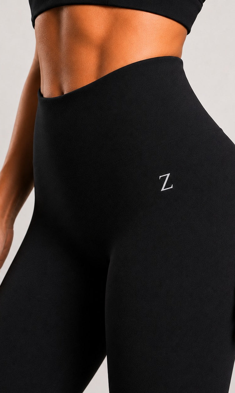 Close-up of a model wearing black high-waisted leggings with a small white embroidered letter Z on the hip, paired with a matching black sports bra, against a light gray background.