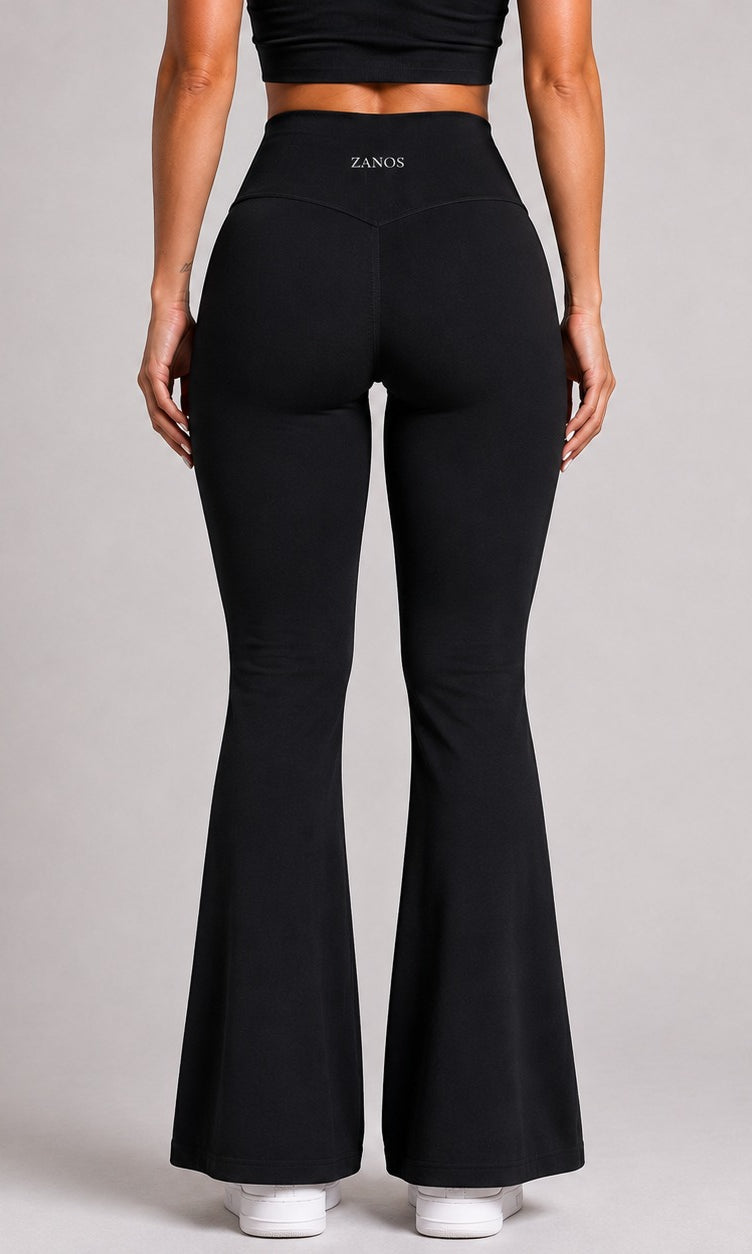 Rear view of a model wearing black high-waisted flared leggings with the brand name ZANOS visible on the waistband, set against a solid light gray studio background.