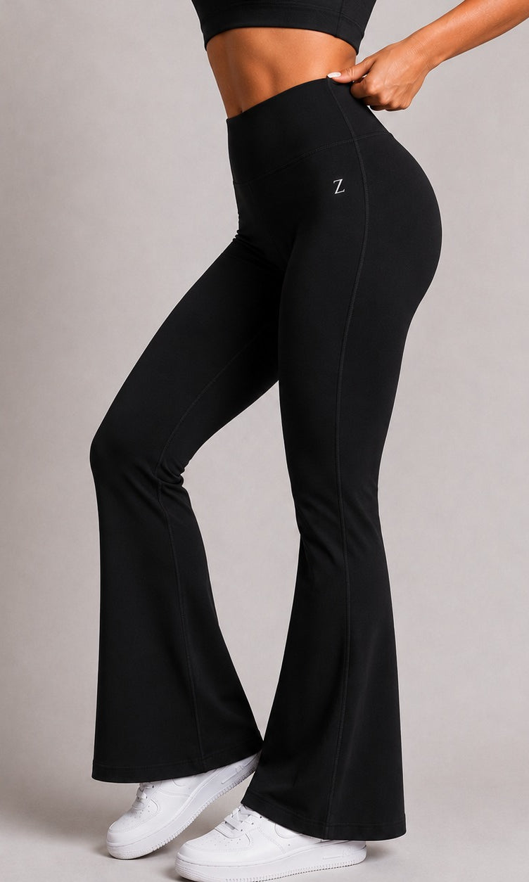 Black high-waisted flared leggings with a small white 'Z' logo on the upper right hip, shown on a model from the waist down against a neutral gray studio background.