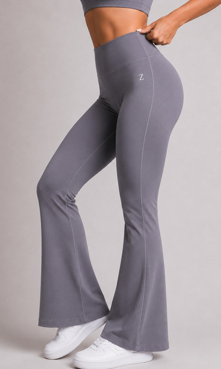 A model wearing high-waisted, flared gray activewear leggings with a small white 'Z' logo on the hip, paired with white sneakers, against a neutral gray studio background.