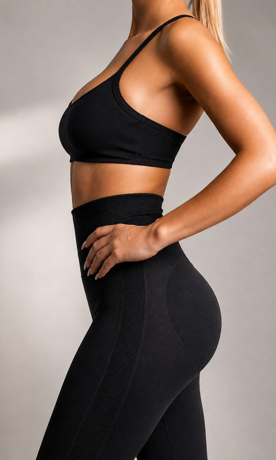 A woman wearing a black sports bra and matching high-waisted black leggings, shown from the side against a neutral gray studio background.