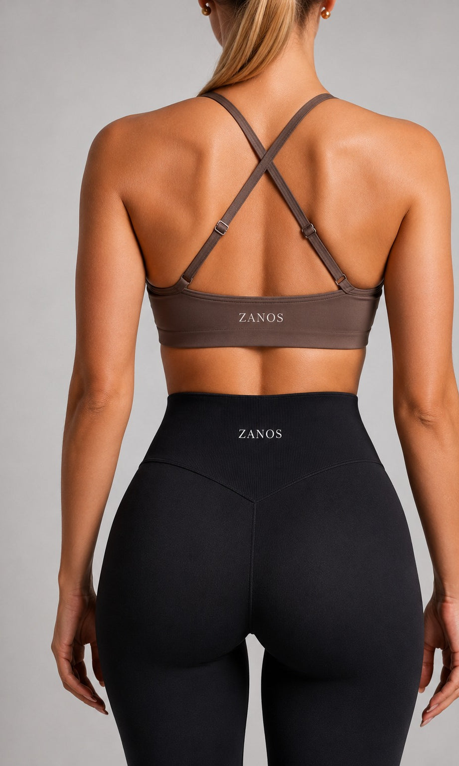 Rear view of a woman wearing a brown ZANOS sports bra with crisscross back straps and black high-waisted leggings, against a neutral gray studio background.