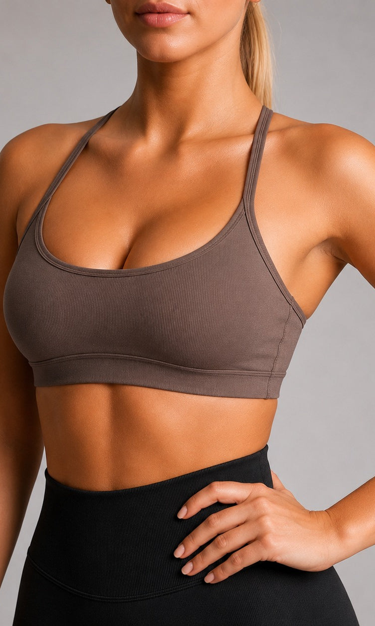 A woman wearing a taupe brown sports bra and black high-waisted leggings, cropped from the chin down, posing against a neutral gray studio background.