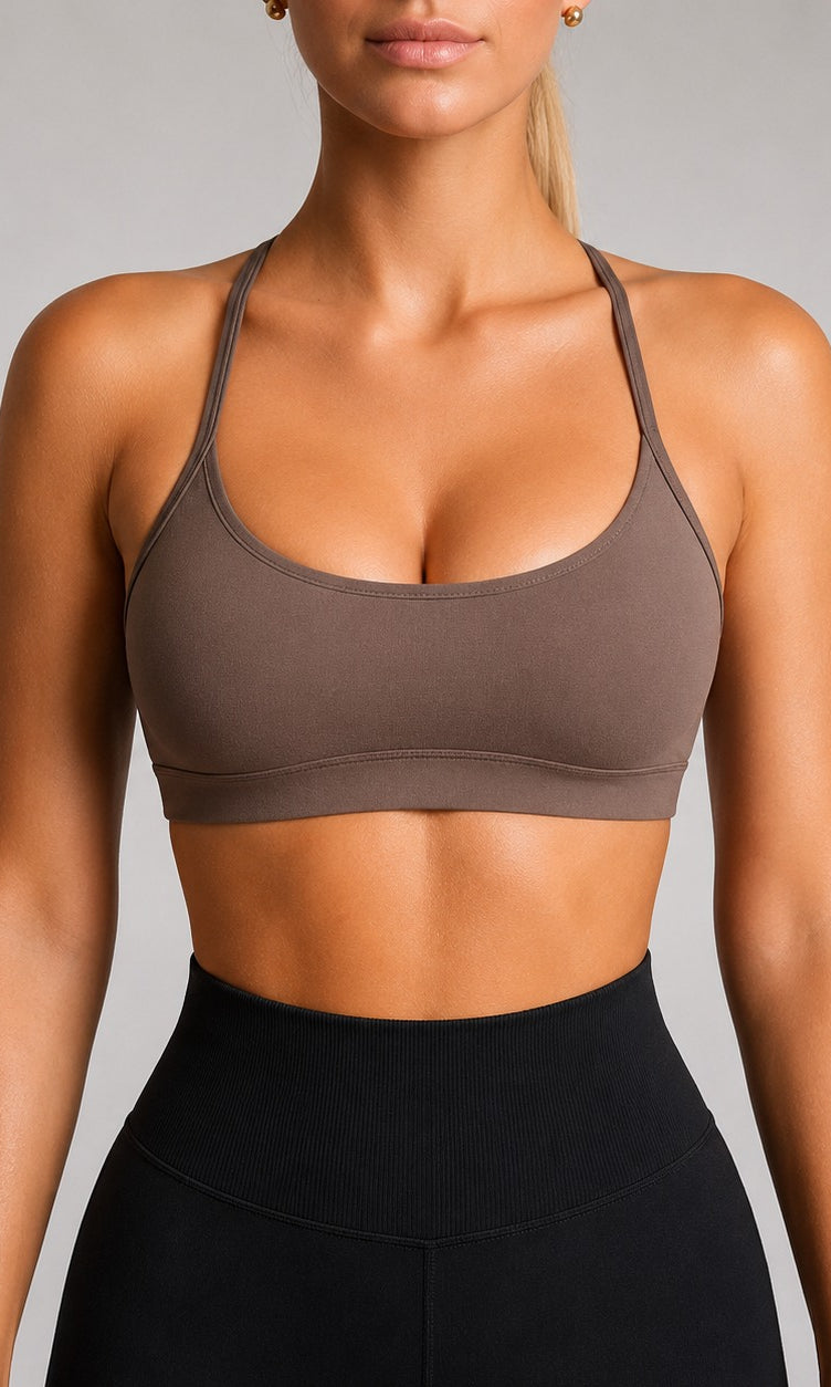 A woman wearing a scoop-neck athletic sports bra in taupe brown, paired with black high-waisted leggings, shown from the lips down against a neutral gray studio background.