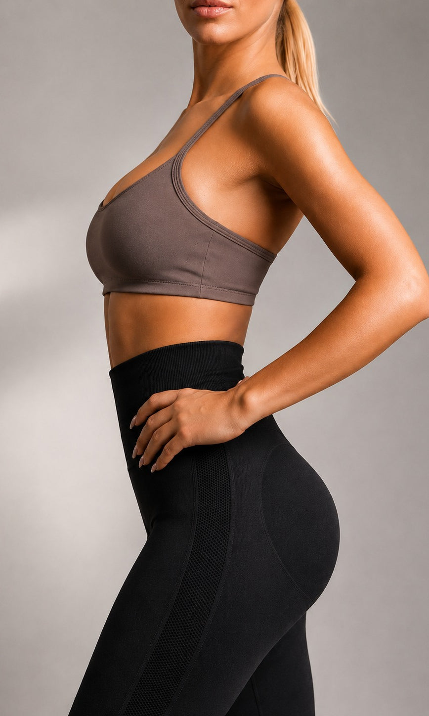 A woman in side profile wearing a taupe-brown sports bra and black high-waisted leggings against a neutral gray studio background.