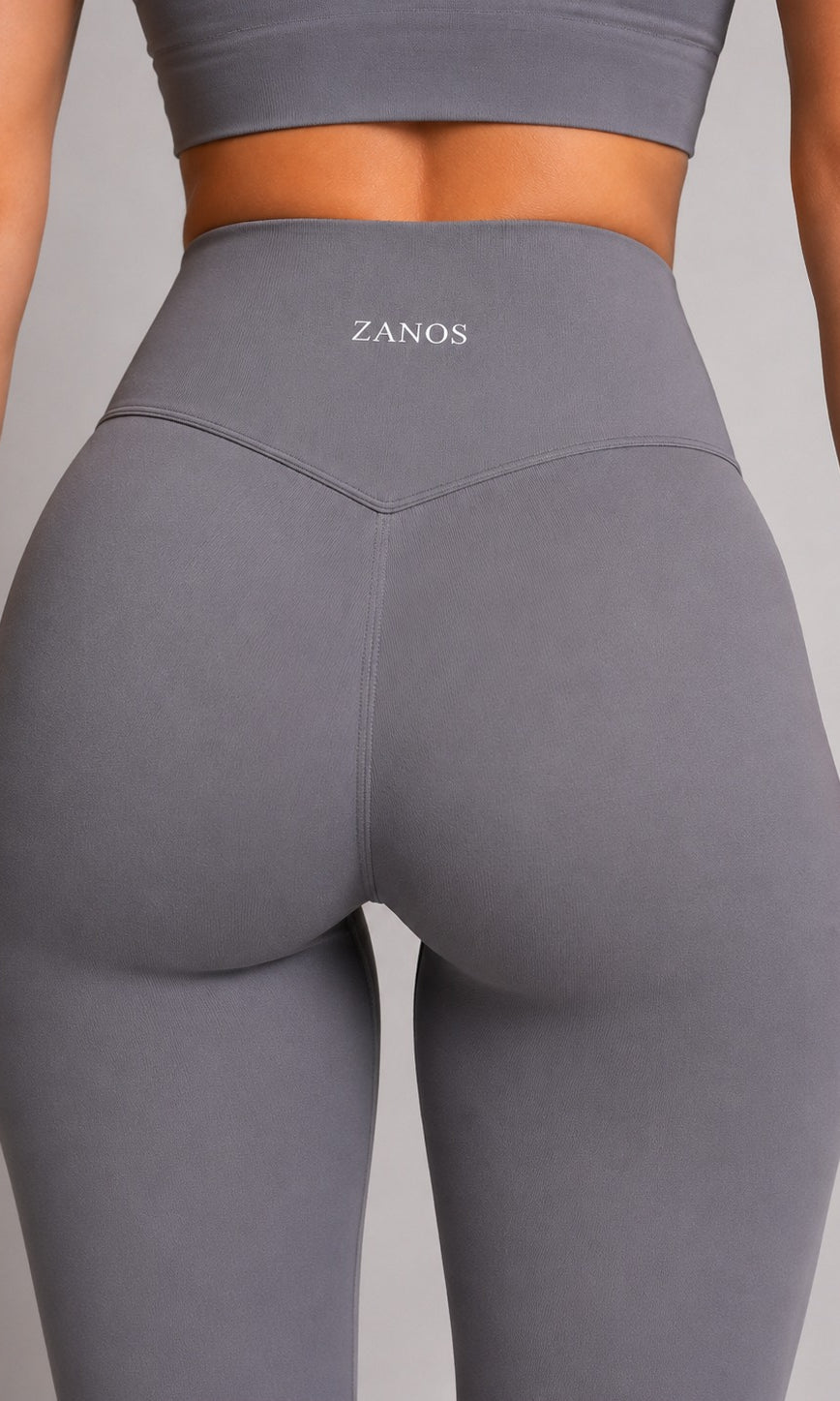 Rear view of a model wearing high-waisted gray ZANOS activewear leggings and a matching sports bra against a neutral gray background.