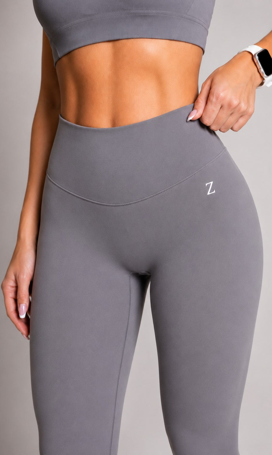 A close-up of a model wearing high-waisted gray athletic leggings with a small white 'Z' logo on the hip, paired with a matching gray sports bra against a neutral gray background.