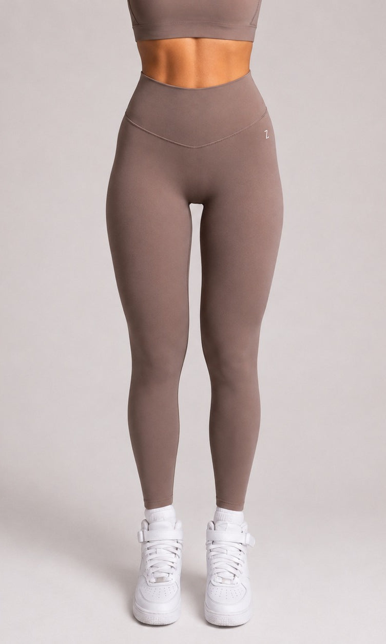 A model from the waist down wearing high-waisted, form-fitting taupe leggings and white sneakers, standing against a solid light gray studio background.