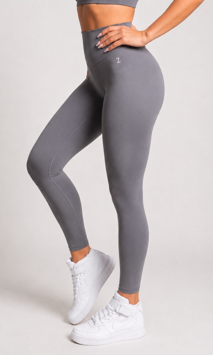A model wearing high-waisted gray leggings with a small white logo on the hip, paired with white sneakers, shown from the waist down against a light gray studio background.