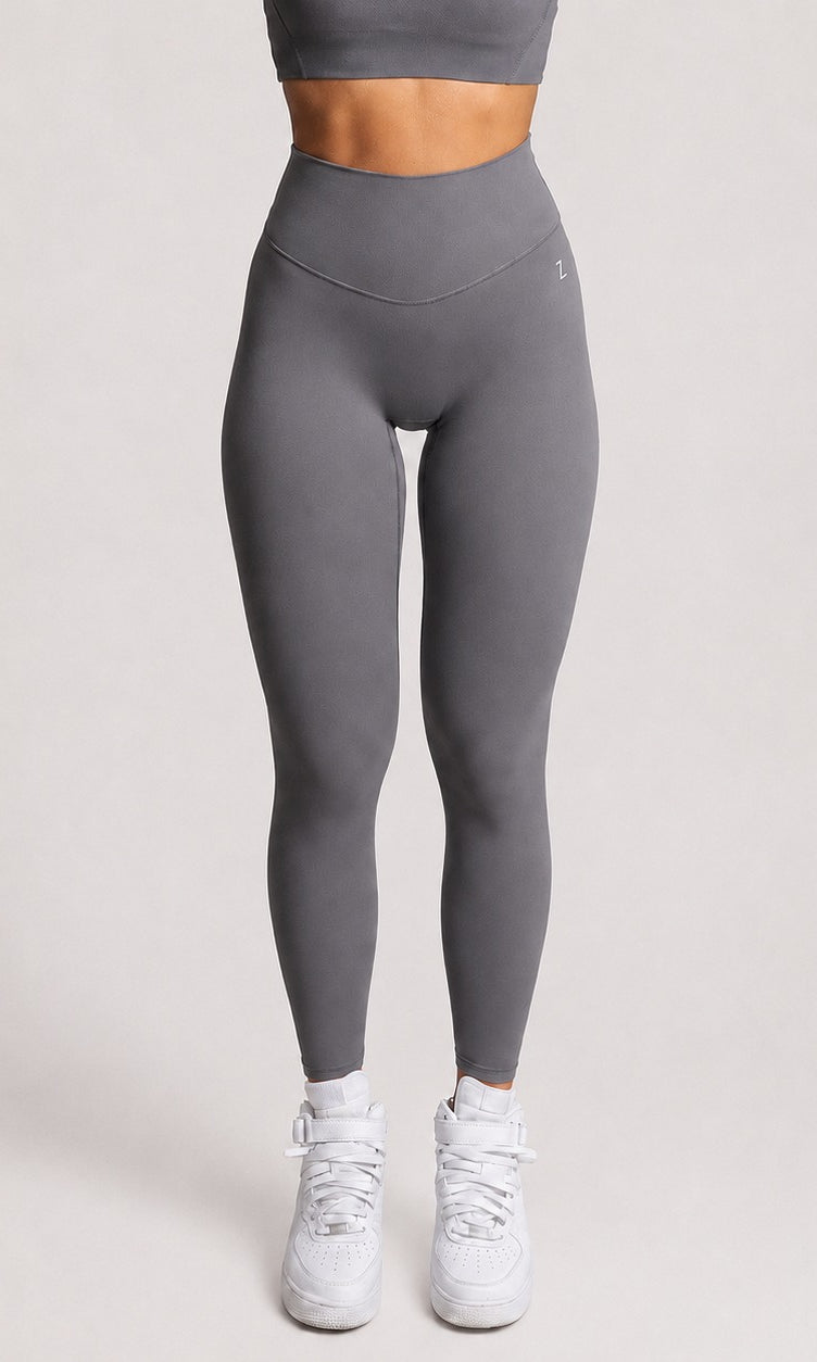 A model from the waist down wearing high-waisted, form-fitting gray leggings and white sneakers, standing against a light gray studio background.