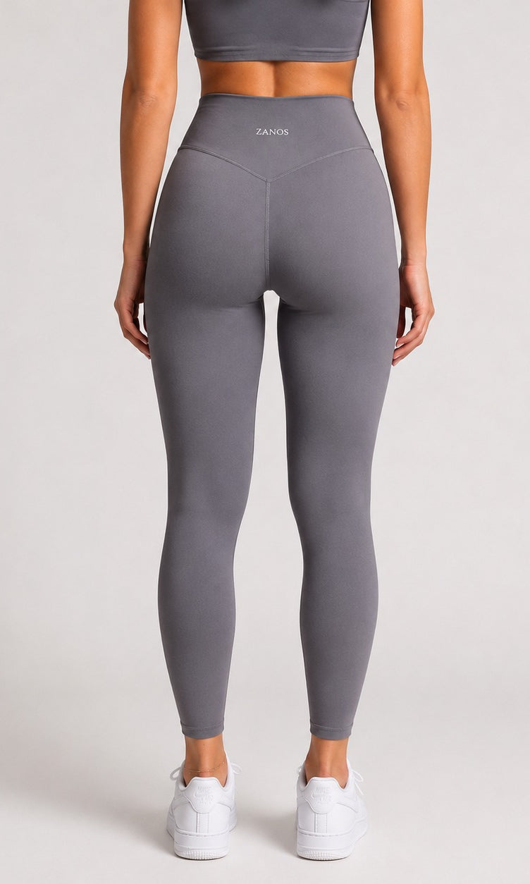 A model shown from the back wearing high-waisted gray leggings with a small white 'ZANOS' logo on the waistband, paired with white sneakers against a light gray studio background.