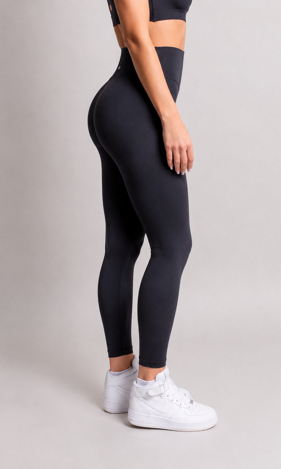 A model wearing high-waisted black leggings and white sneakers, shown from a side-back angle against a neutral light gray studio background.
