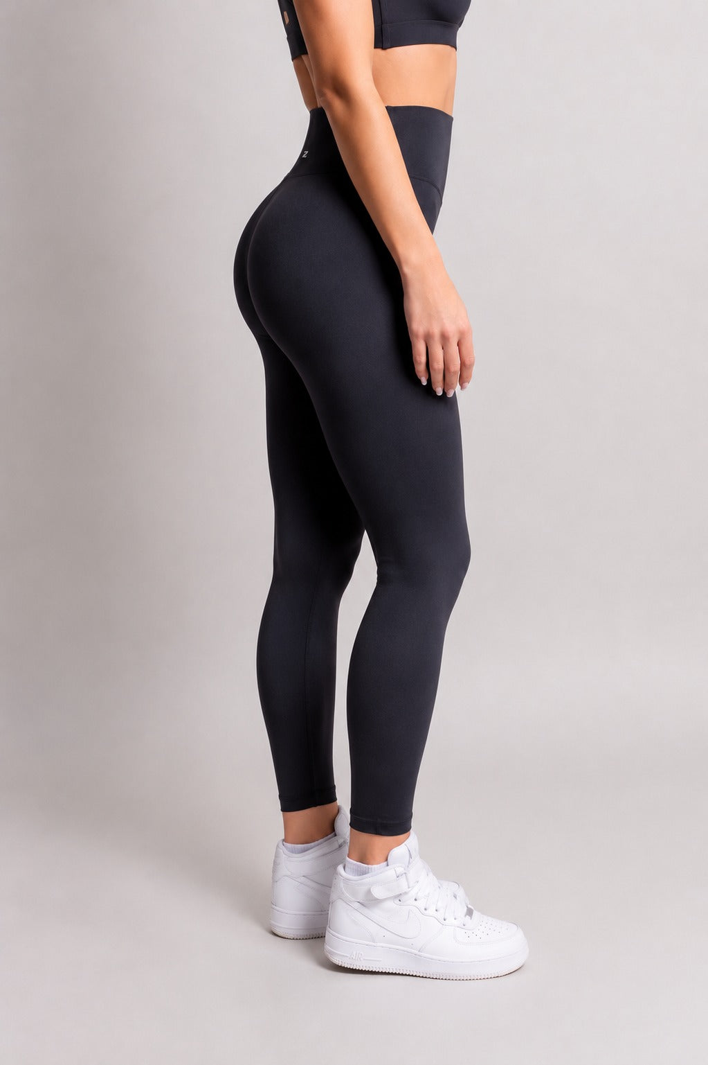 A model wearing high-waisted black leggings and white sneakers, shown from a side-back angle against a neutral light gray studio background.
