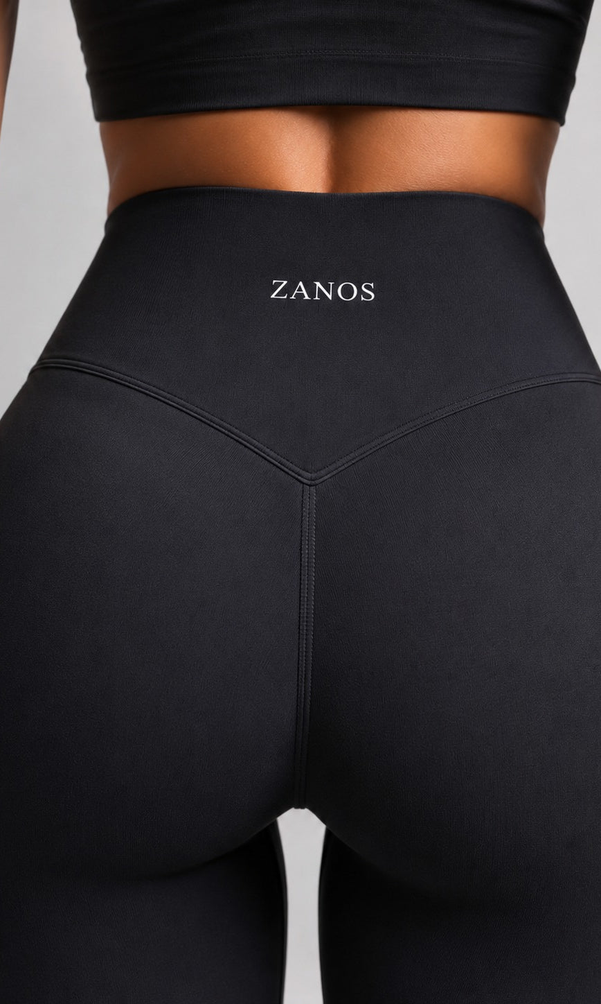 Close-up back view of a model wearing black high-waisted leggings with the brand name ZANOS printed in white on the waistband, set against a solid gray background.
