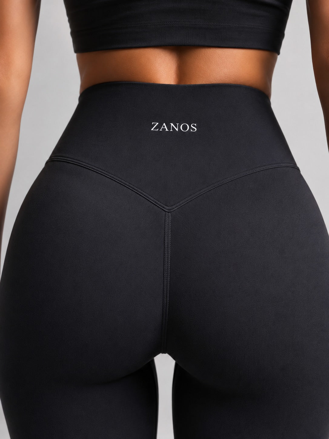 Close-up back view of a model wearing black high-waisted leggings with the brand name ZANOS printed in white on the waistband, set against a solid gray background.
