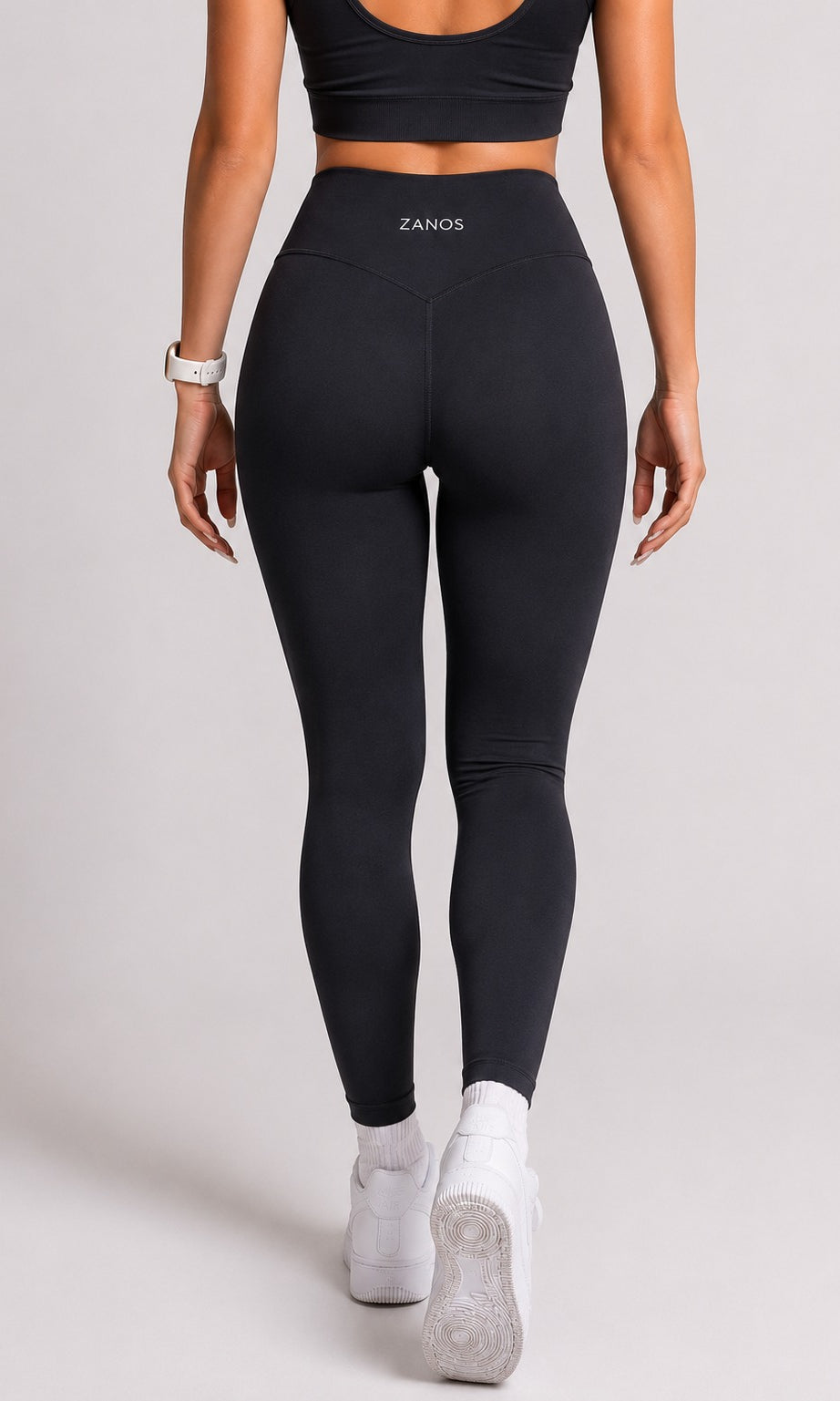 Rear view of a woman wearing black high-waisted ZANOS leggings, a black sports bra, and white sneakers against a light gray studio background.