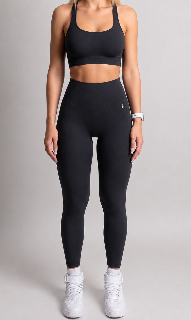 A woman modeling a black activewear set consisting of a sports bra and high-waisted leggings, paired with white sneakers, against a neutral gray studio background.
