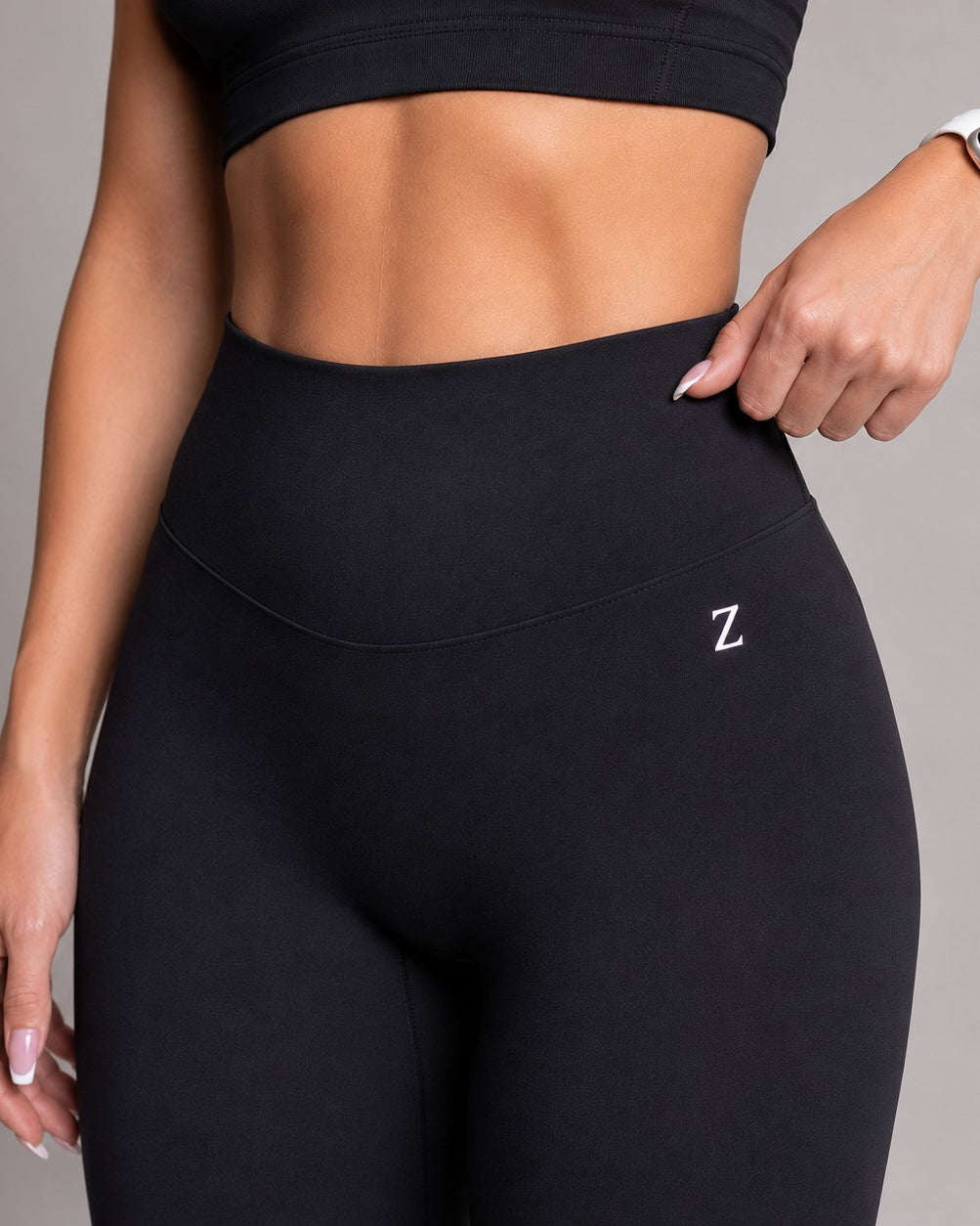 A close-up of a model wearing black high-waisted athletic leggings with a small white embroidered 'Z' logo on the hip, paired with a matching black sports bra. The model is shown from the chest down, with one hand pulling slightly at the side of the waistband to demonstrate the stretch.
