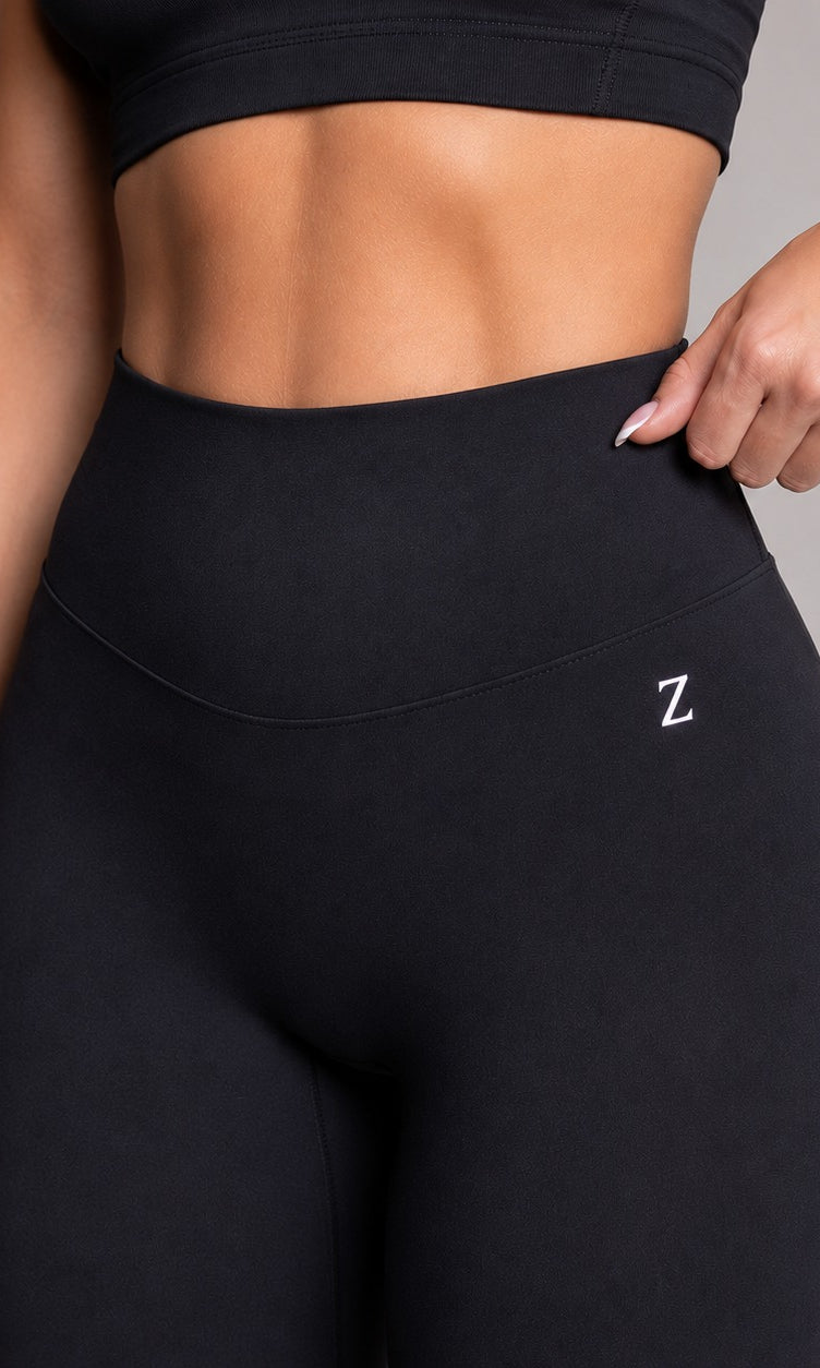 A close-up of a model wearing black high-waisted athletic leggings with a small white embroidered 'Z' logo on the hip, paired with a matching black sports bra. The model is shown from the chest down, with one hand pulling slightly at the side of the waistband to demonstrate the stretch.