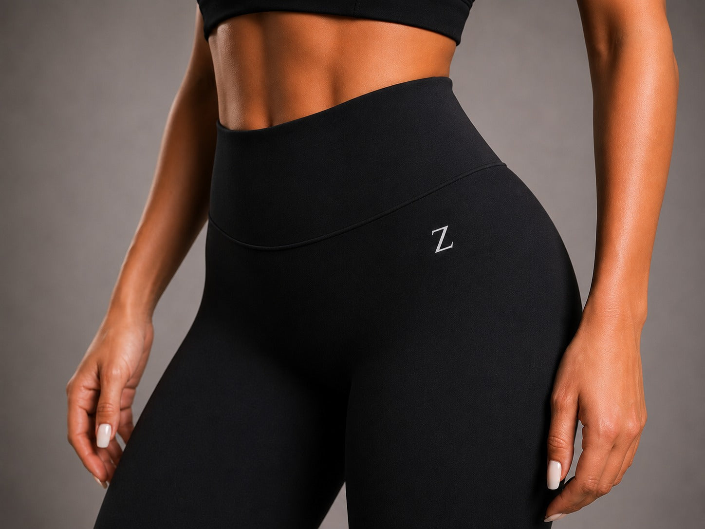 Close-up of a model wearing black high-waisted leggings with a small white embroidered 'Z' logo on the hip, paired with a matching black sports bra, against a neutral gray studio background.