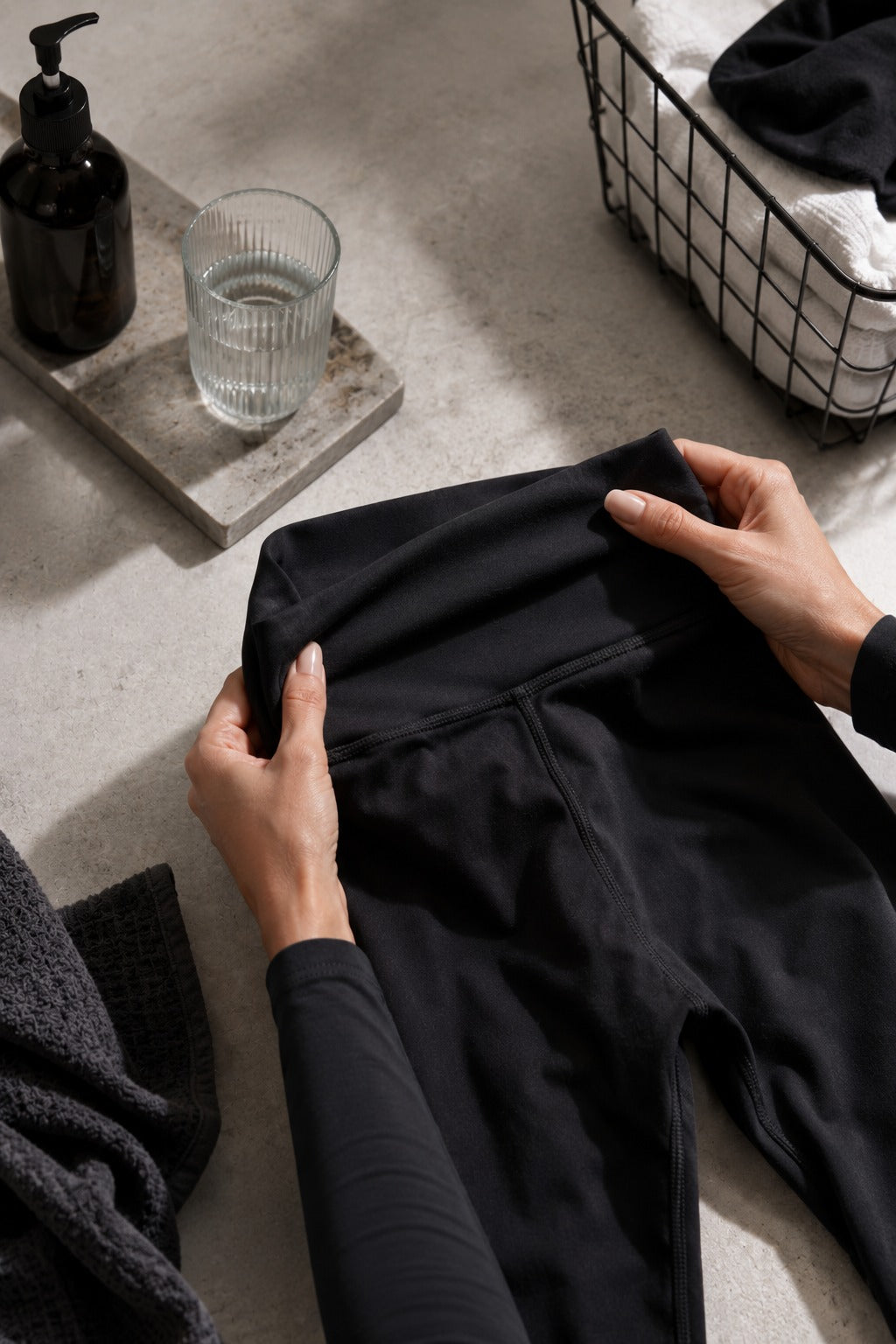A person's hands holding up a pair of black leggings to show the high-waisted waistband, set in a styled home environment with a glass of water, a soap dispenser, and a laundry basket.