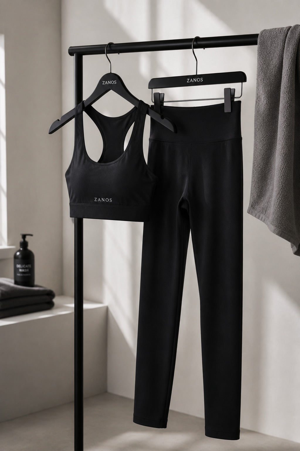 Black activewear set from ZANOS, including a racerback sports bra and high-waisted leggings, displayed on black hangers hanging from a clothing rack in a minimalist, sunlit room.
