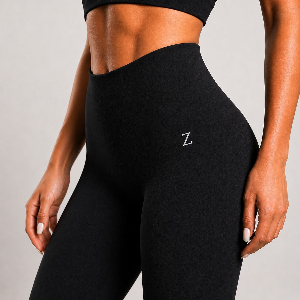 Close-up of a model wearing black high-waisted leggings with a small white embroidered letter Z on the hip, paired with a matching black sports bra, against a light gray background.
