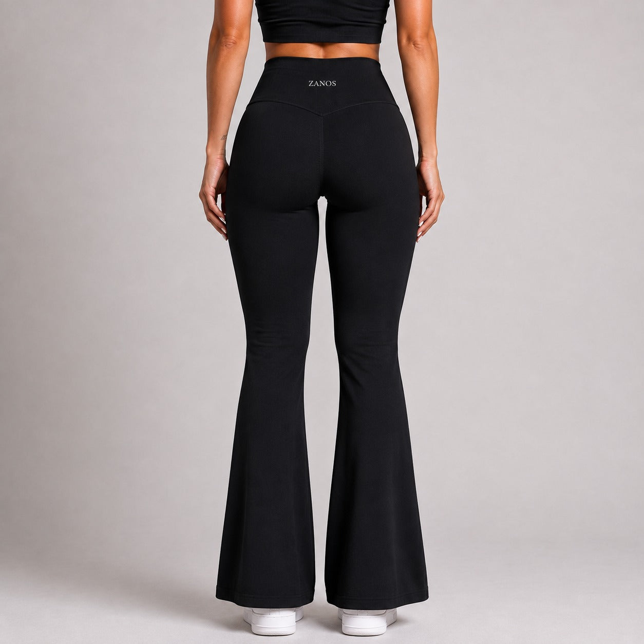 Rear view of a model wearing black high-waisted flared leggings with the brand name ZANOS visible on the waistband, set against a solid light gray studio background.