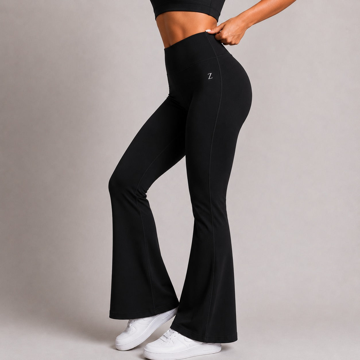 Black high-waisted flared leggings with a small white 'Z' logo on the upper right hip, shown on a model from the waist down against a neutral gray studio background.