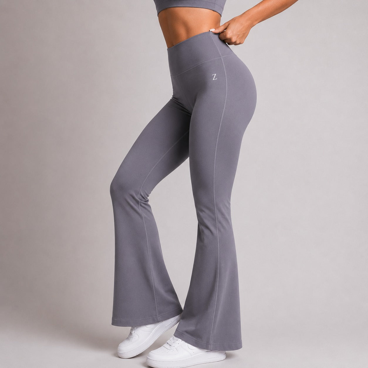 A model wearing high-waisted, flared gray activewear leggings with a small white 'Z' logo on the hip, paired with white sneakers, against a neutral gray studio background.