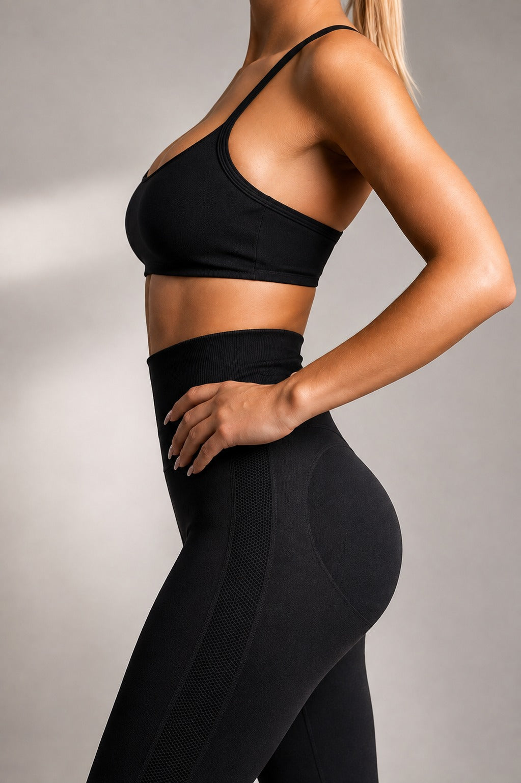 A woman wearing a black sports bra and matching high-waisted black leggings, shown from the side against a neutral gray studio background.