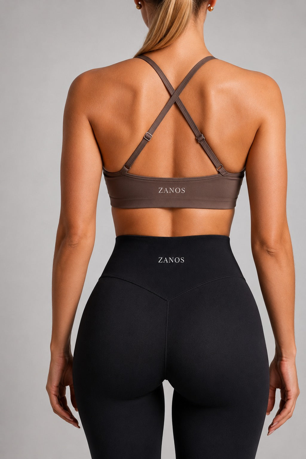 Rear view of a woman wearing a brown ZANOS sports bra with crisscross back straps and black high-waisted leggings, against a neutral gray studio background.