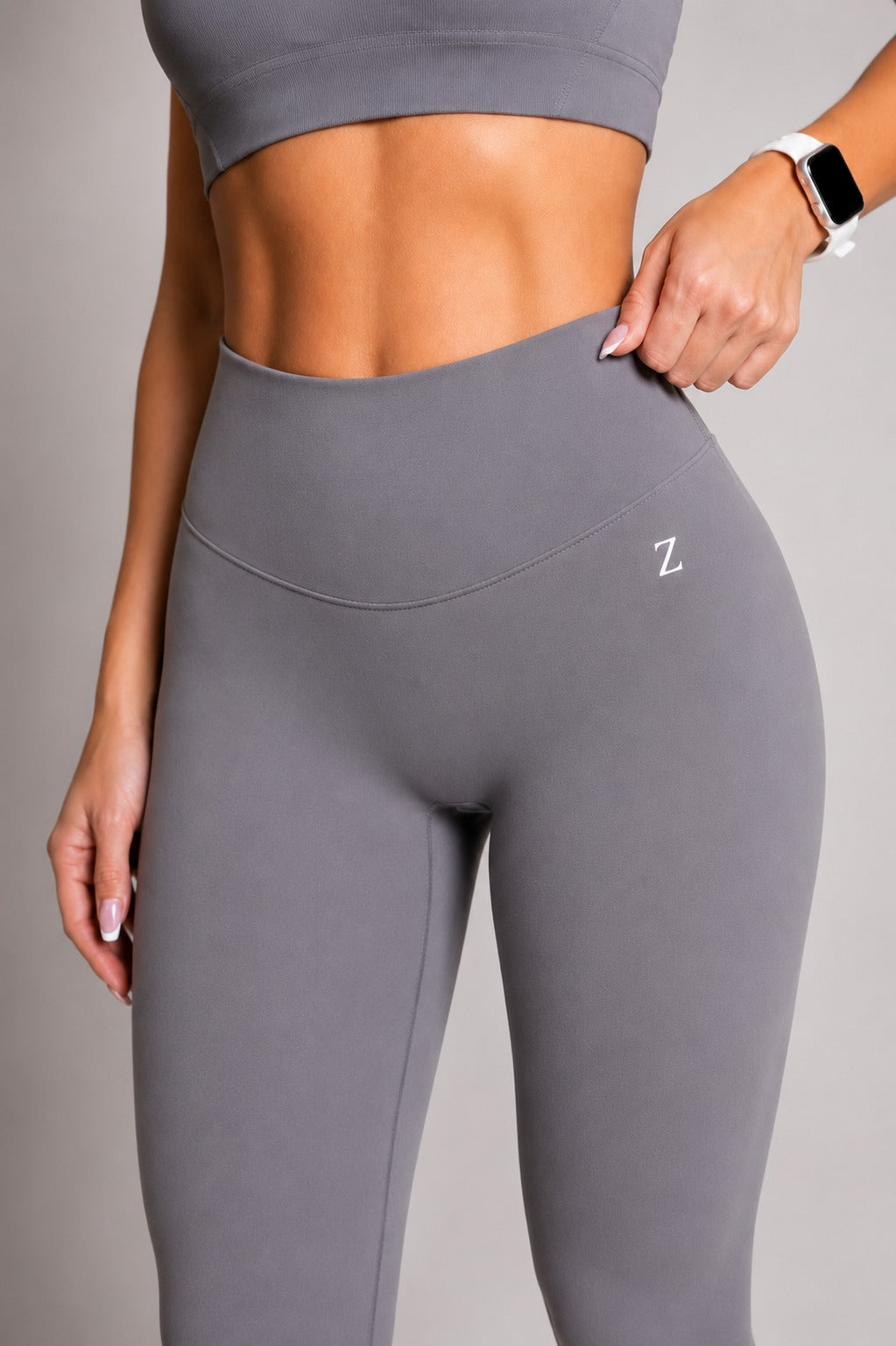 A close-up of a model wearing high-waisted gray athletic leggings with a small white 'Z' logo on the hip, paired with a matching gray sports bra against a neutral gray background.