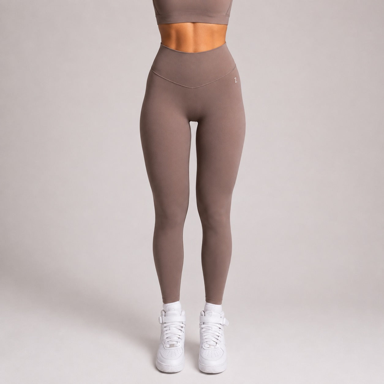 A model from the waist down wearing high-waisted, form-fitting taupe leggings and white sneakers, standing against a solid light gray studio background.