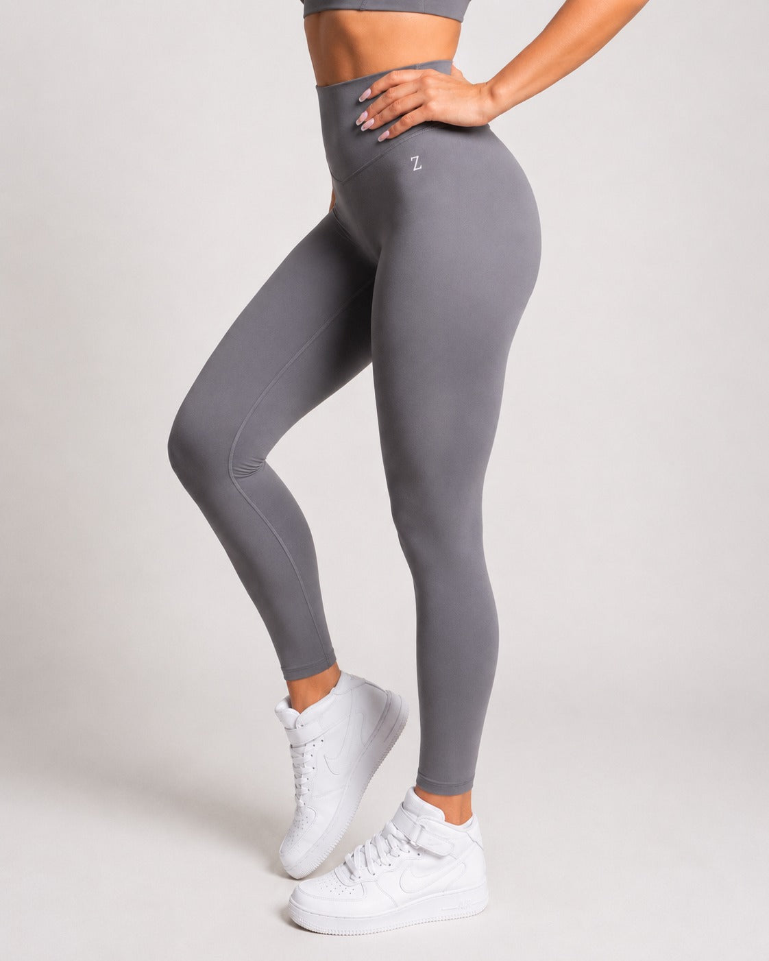 A model wearing high-waisted gray leggings with a small white logo on the hip, paired with white sneakers, shown from the waist down against a light gray studio background.