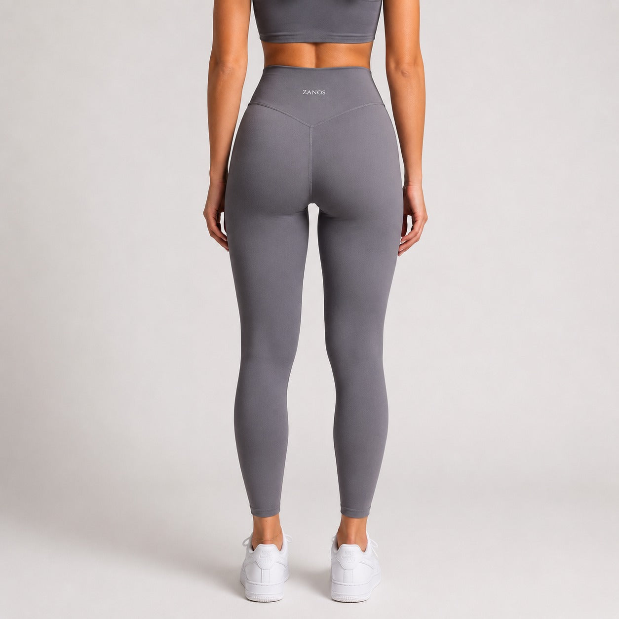 A model shown from the back wearing high-waisted gray leggings with a small white 'ZANOS' logo on the waistband, paired with white sneakers against a light gray studio background.