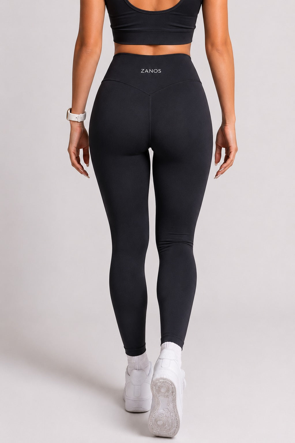 Rear view of a woman wearing black high-waisted ZANOS leggings, a black sports bra, and white sneakers against a light gray studio background.