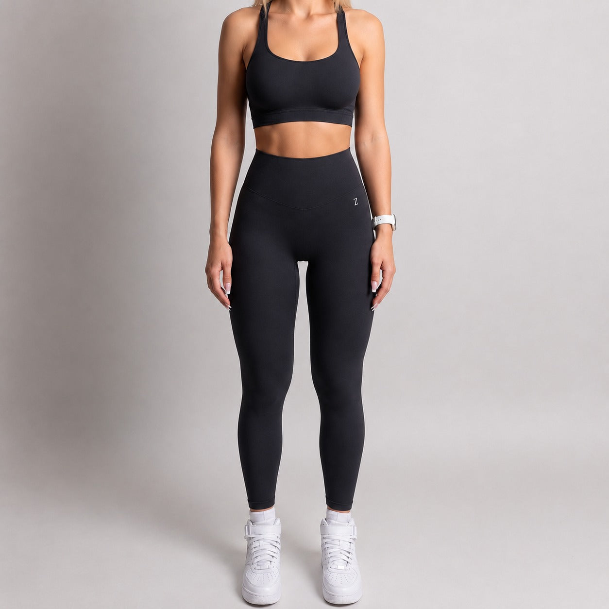 A woman modeling a black activewear set consisting of a sports bra and high-waisted leggings, paired with white sneakers, against a neutral gray studio background.